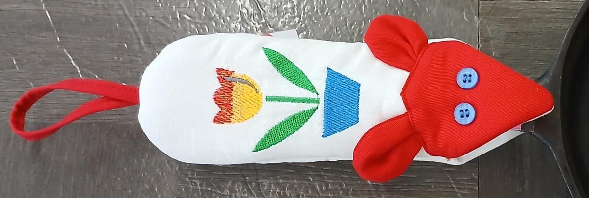 A white fabric mouse-shaped skiller handle potholder with a red loop handle, red ears, blue button eyes, and an embroidered tulip flower design on the back, shown on a gray wood-textured surface.
