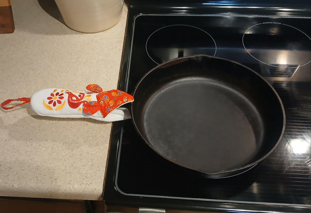 A black cast iron skillet sits on a black electric stovetop, with a white handle potholder featuring a red, orange, and yellow floral pattern resting on the beige countertop beside it.