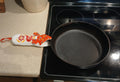 A black cast iron skillet sits on a black electric stovetop, with a white handle potholder featuring a red, orange, and yellow floral pattern resting on the beige countertop beside it.