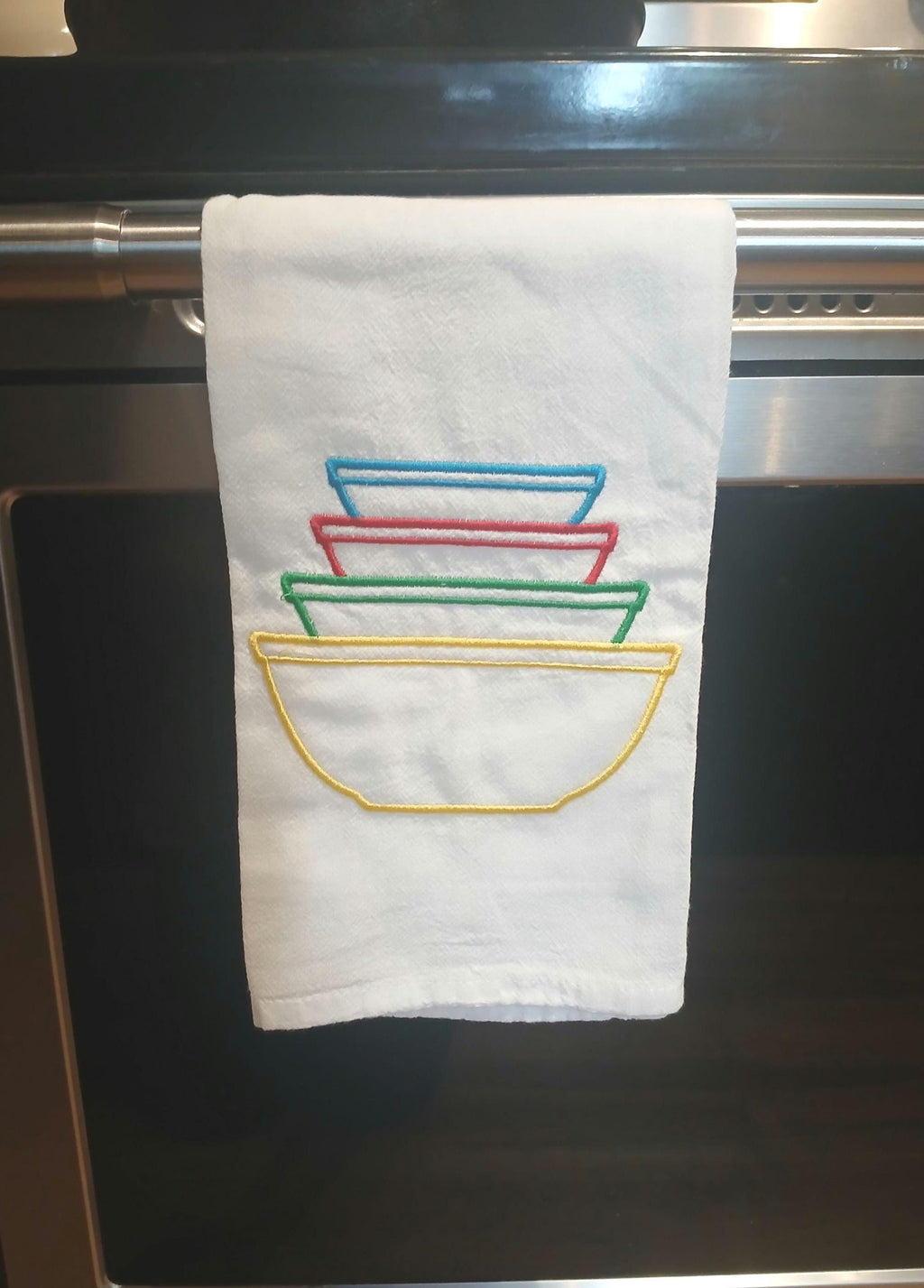 A white kitchen towel hanging from a metal oven handle, featuring embroidered stacked bowls in blue, red, green, and yellow.