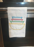A white kitchen towel hanging from a metal oven handle, featuring embroidered stacked bowls in blue, red, green, and yellow.