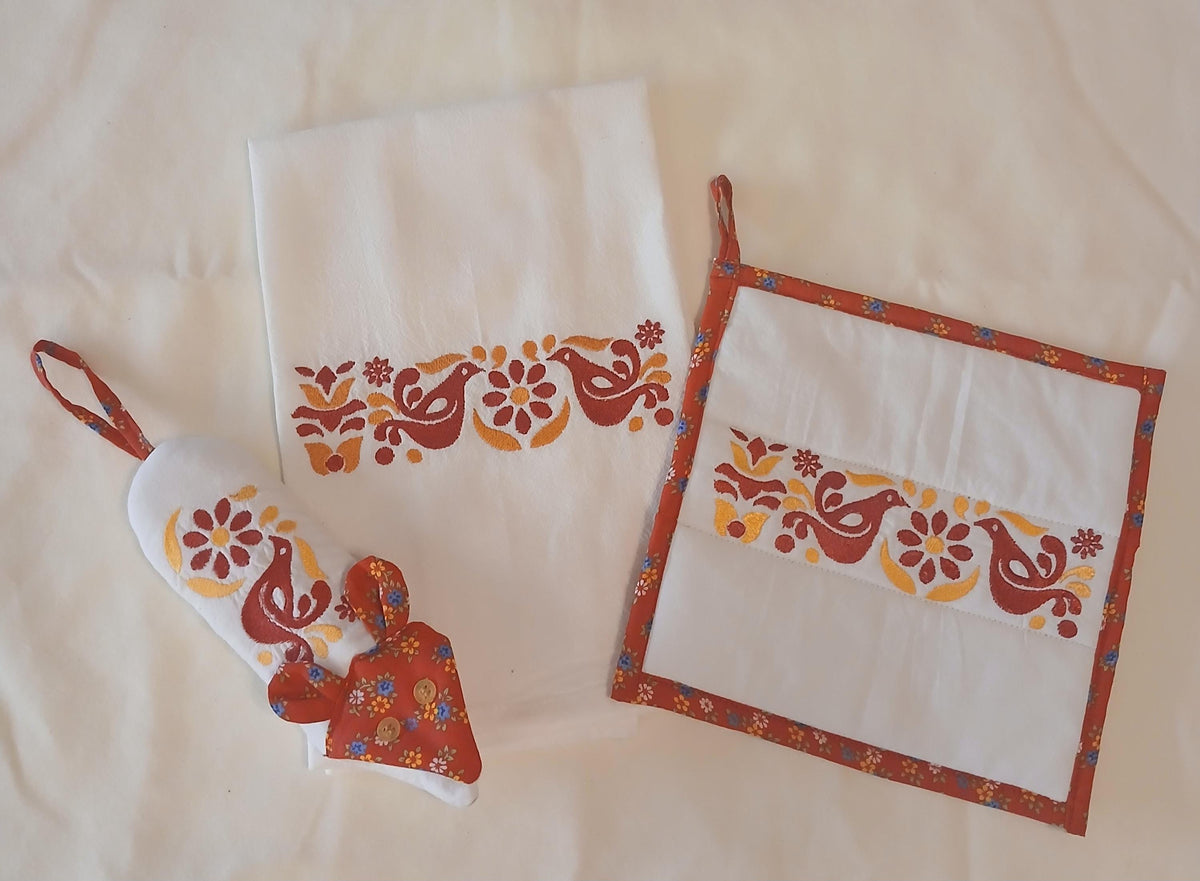 A coordinated kitchen textile set laid flat on a cream fabric background, including a white flour sack towel with a red and orange floral-bird motif, a matching square potholder with a red border and hanging loop, and a handle potholder with the same motif and a red face with button eyes and fabric ears.