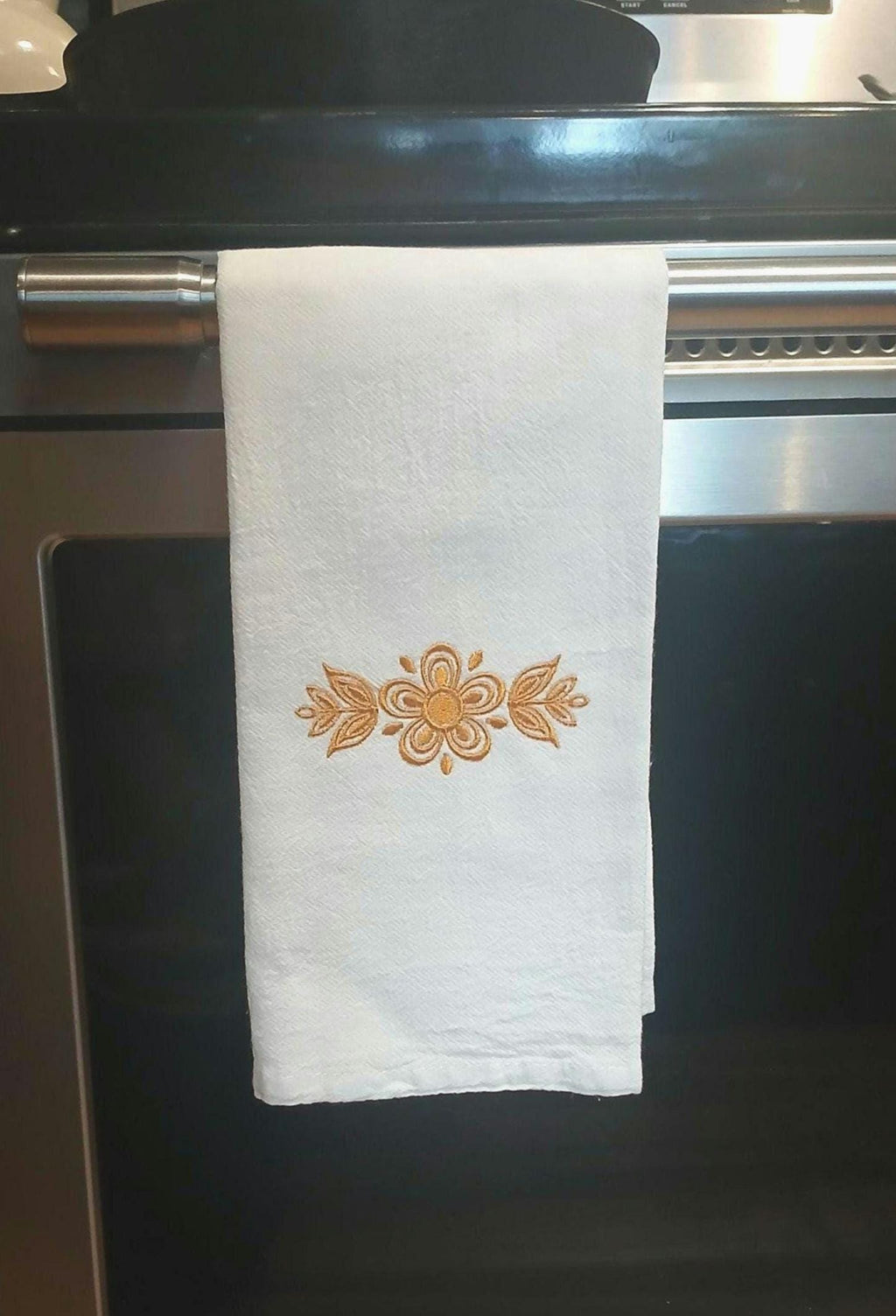 A white kitchen towel with an embroidered gold floral design hanging from a stainless steel oven handle.