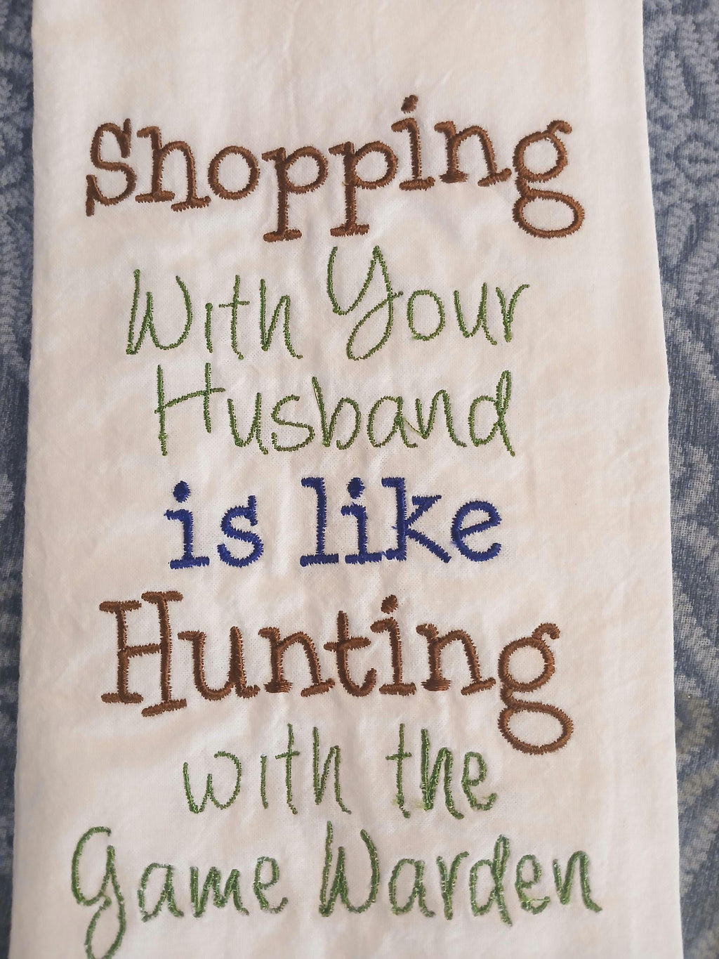 A close-up of a white embroidered kitchen towel featuring the humorous phrase 'Shopping With Your Husband is like Hunting with the Game Warden' stitched in brown, green, and blue thread.