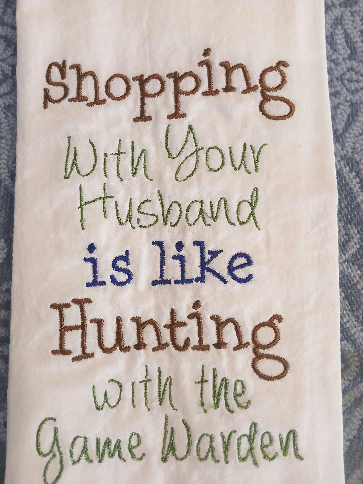 A close-up of a white embroidered kitchen towel featuring the humorous phrase 'Shopping With Your Husband is like Hunting with the Game Warden' stitched in brown, green, and blue thread.