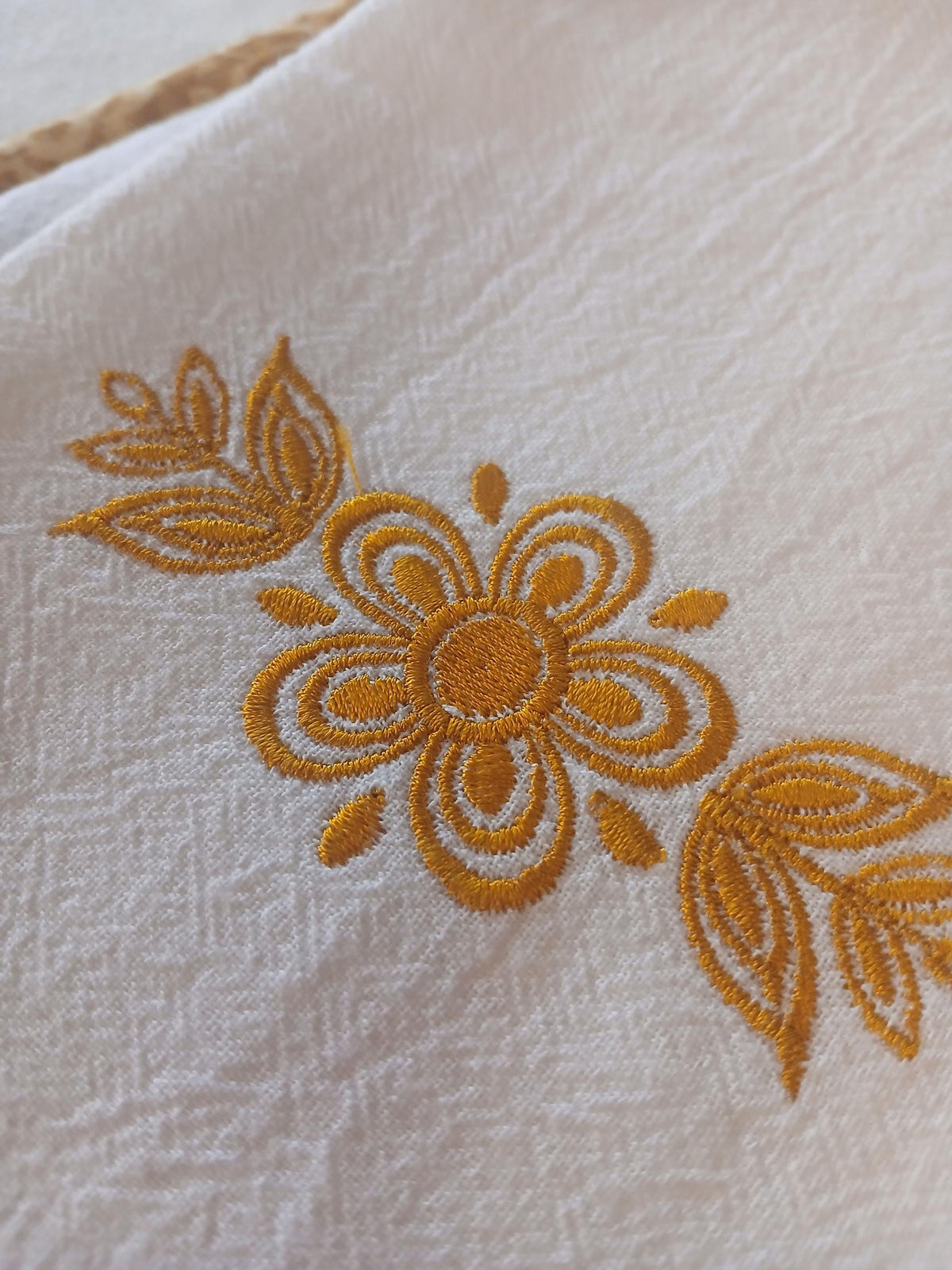 A close-up of a white flour sack towel featuring a decorative gold embroidered floral pattern inspired by Pyrex Butterfly Gold.