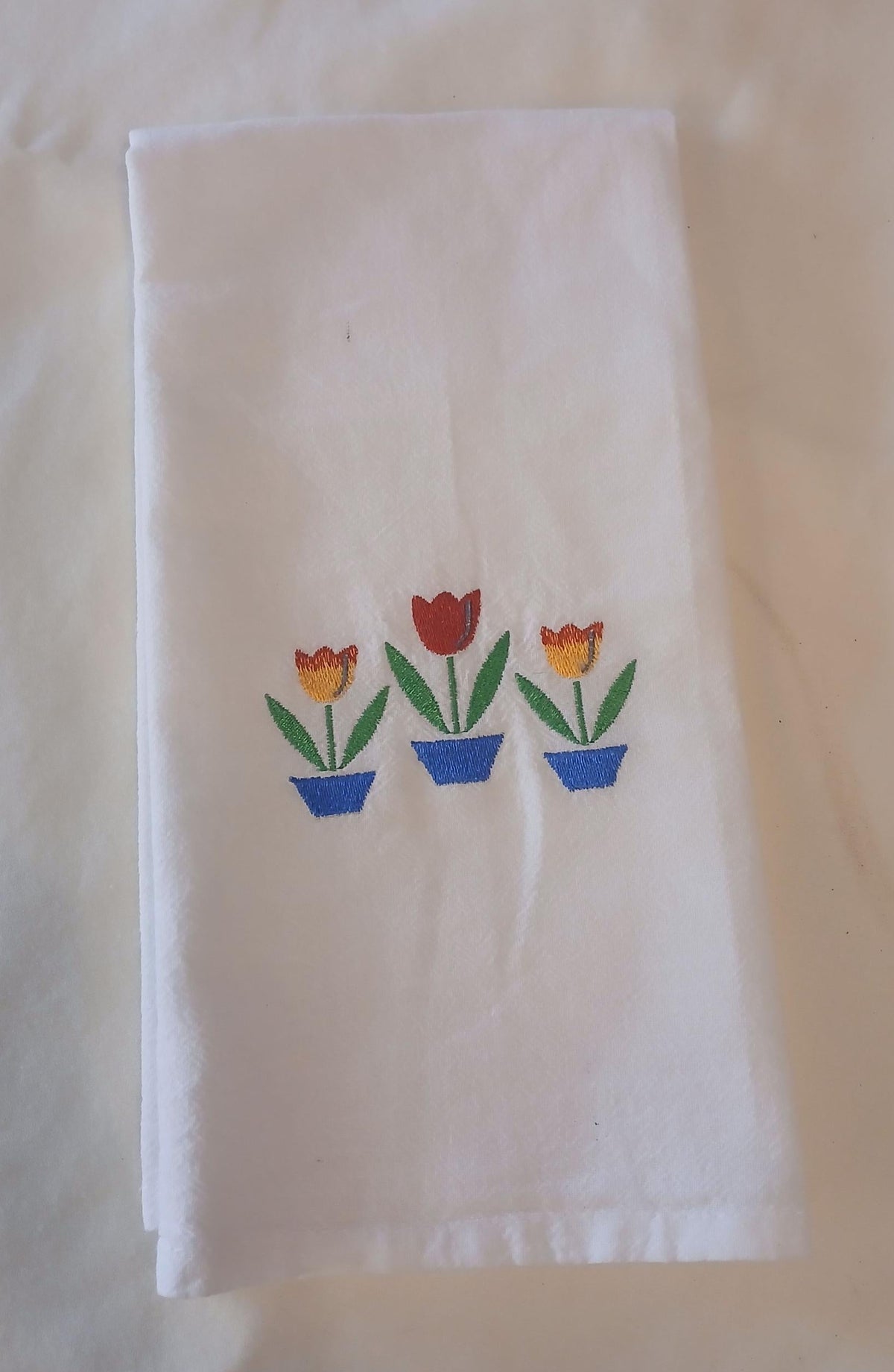A folded white cotton kitchen towel featuring three embroidered potted tulips in blue pots, displayed on an off-white fabric background.