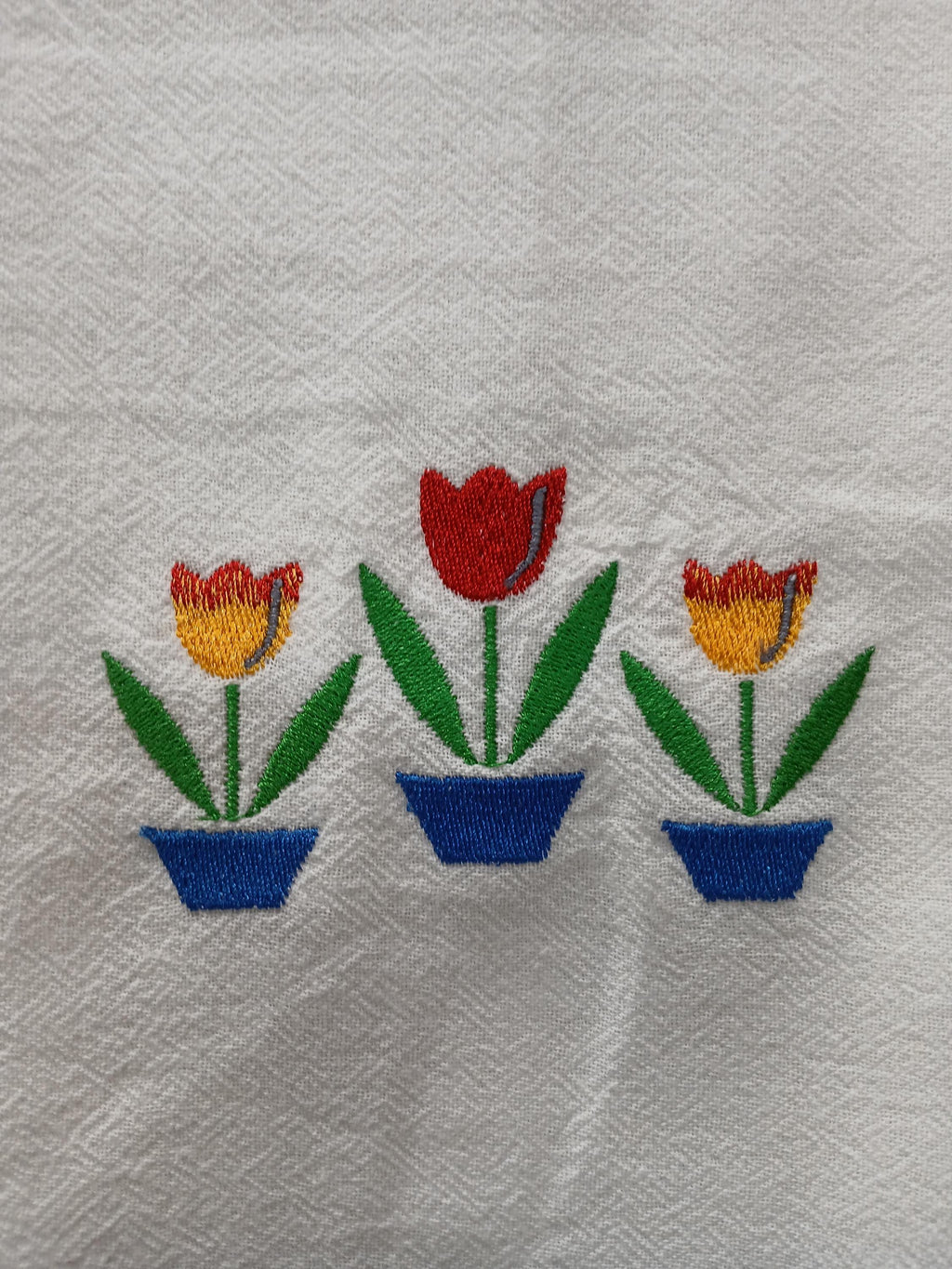 A close-up of a white textured fabric, likely a napkin or table linen, featuring an embroidered motif of three potted tulips in blue pots.