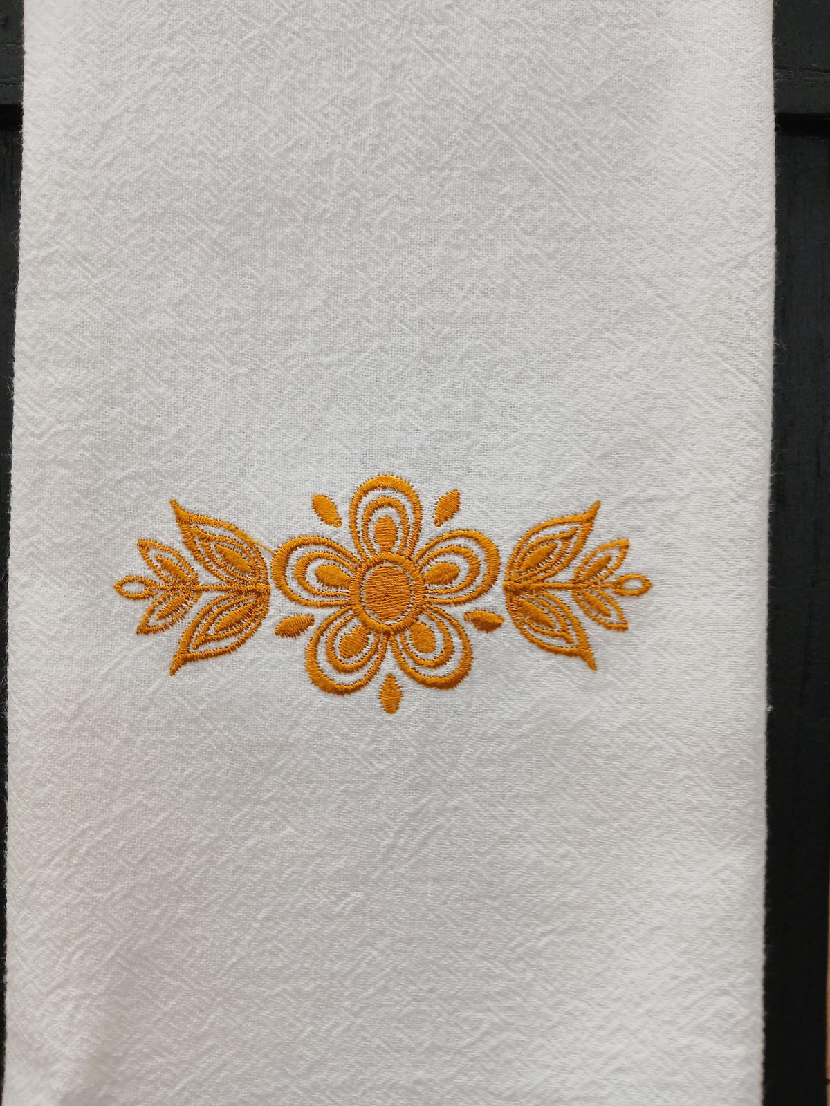 A close-up of a white textured kitchen towel, featuring a central gold embroidered floral and leaf motif.