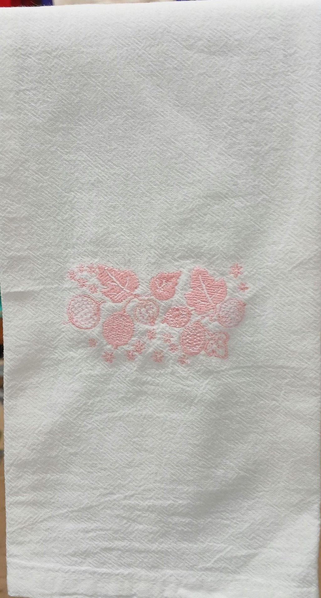 A white cotton kitchen towel with a pink embroidered floral pattern in the center, hanging on a dark rack.