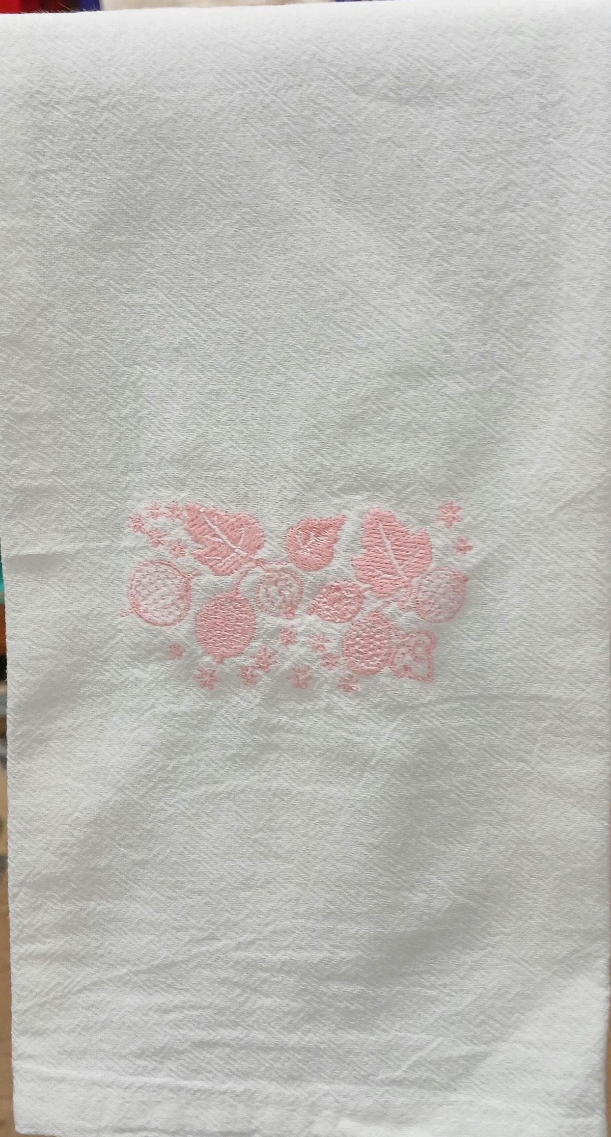 A white cotton kitchen towel with a pink embroidered floral pattern in the center, hanging on a dark rack.