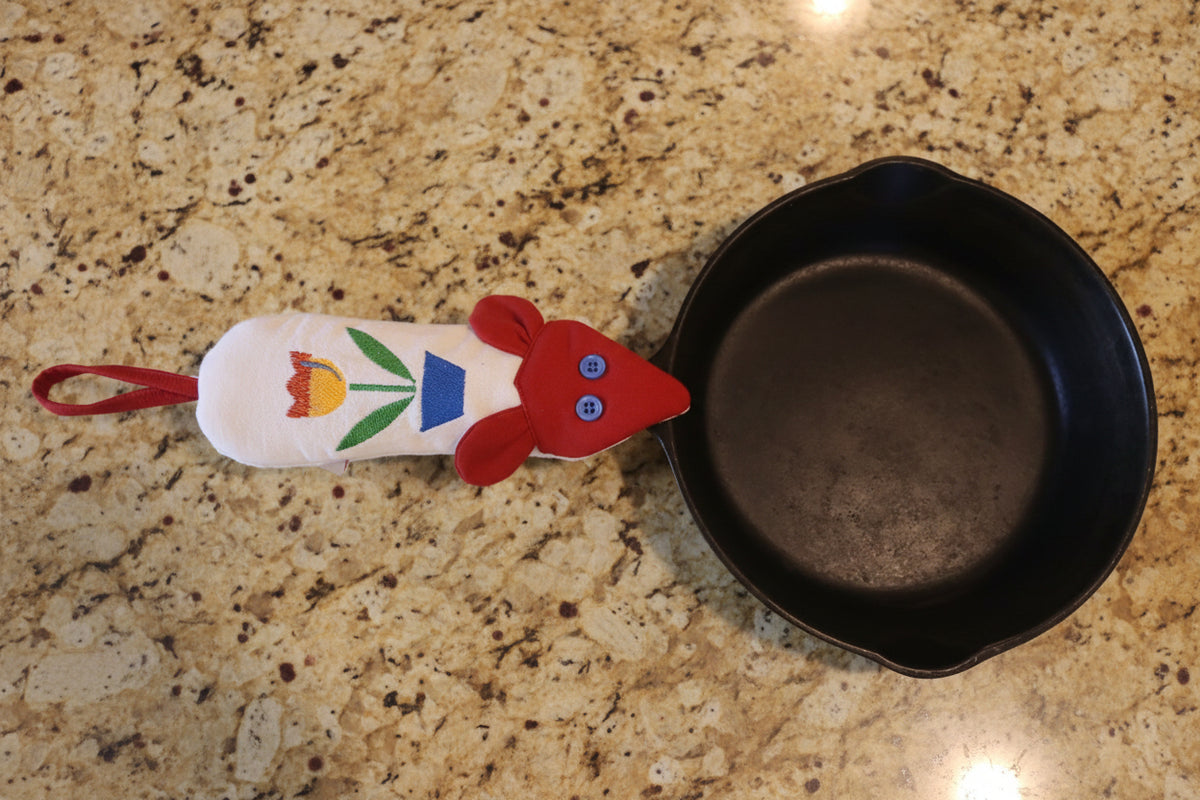 A black cast iron skillet with a white fabric handle potholder featuring a red face, blue button eyes and red ears, and a red and orange tulip design, resting on a cream-colored fabric background.