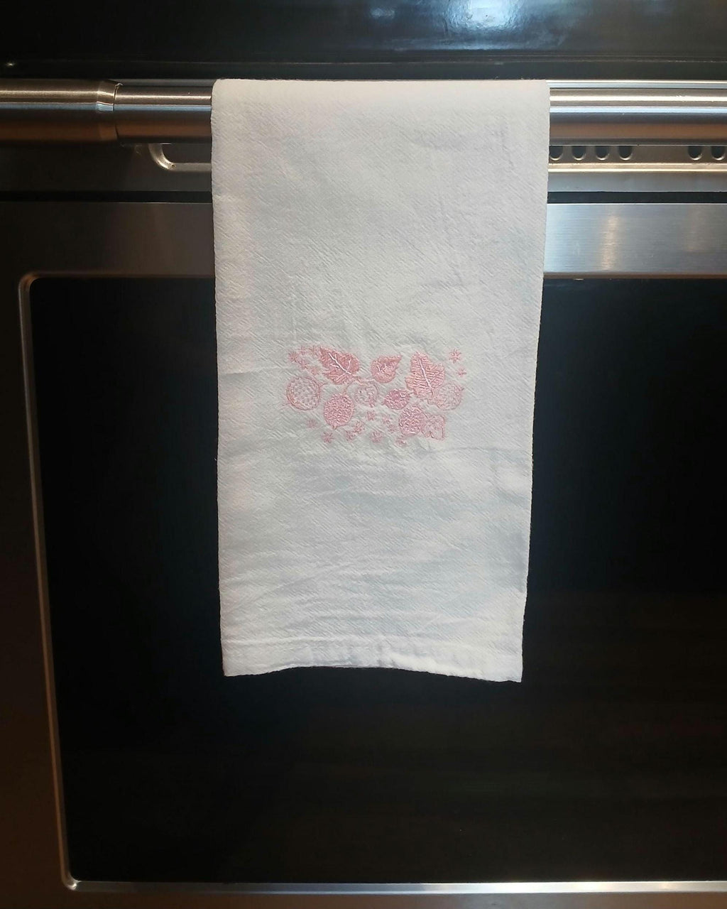 A white kitchen towel with a pink floral pattern in the center, hanging from a stainless steel oven handle.
