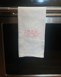 A white kitchen towel with a pink floral pattern in the center, hanging from a stainless steel oven handle.