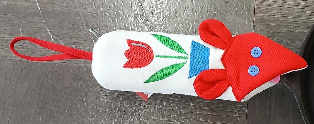 A handmade fabric mouse-shaped skillet handle potholder with a red body, red ears, blue button eyes, a green and red tulip design, and a red hanging loop, shown on a gray wood table.