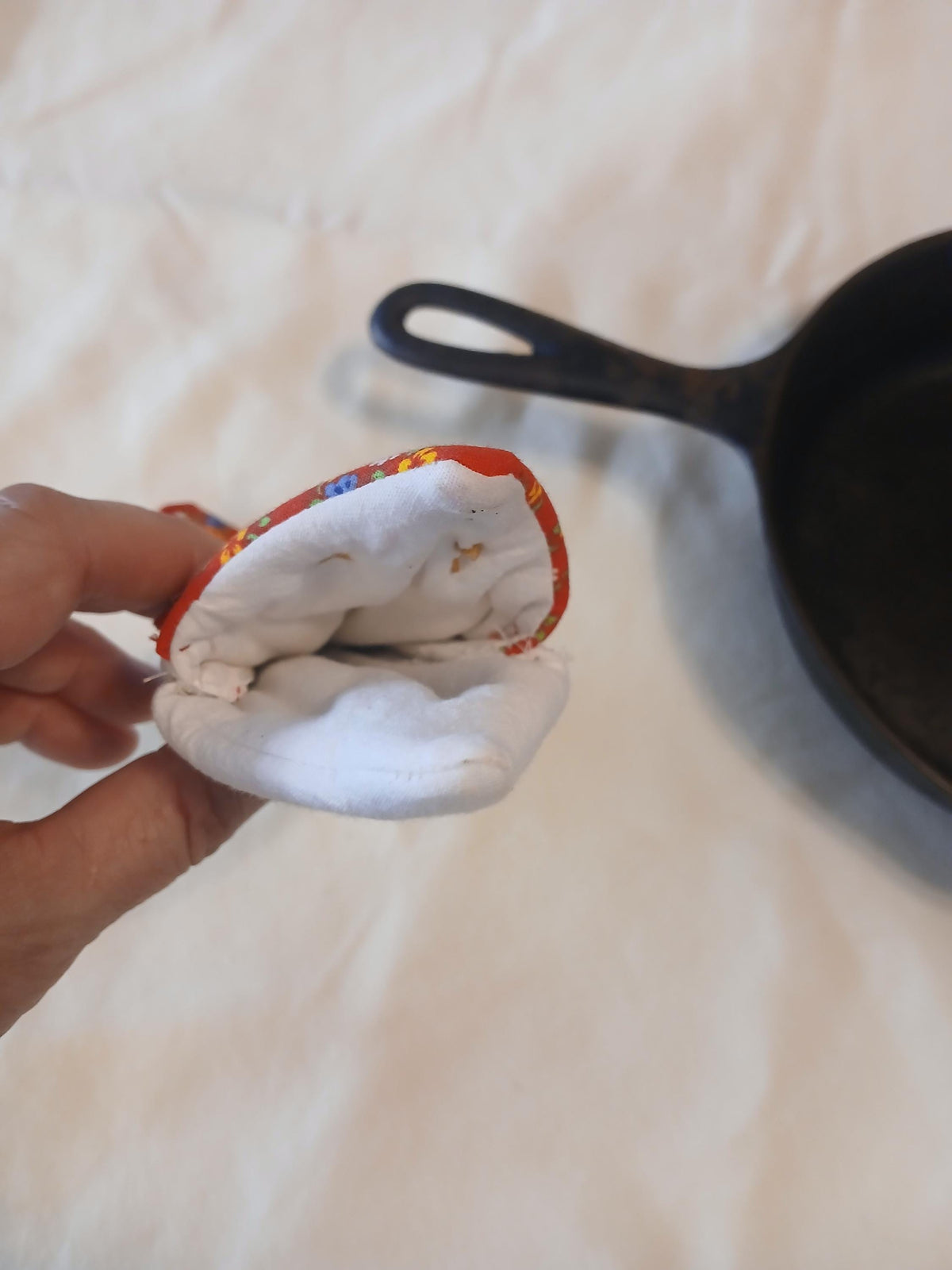 A hand holding a small fabric oven mitt with a colorful patterned edge and white interior, with a black cast iron skillet in the background on a light fabric surface.