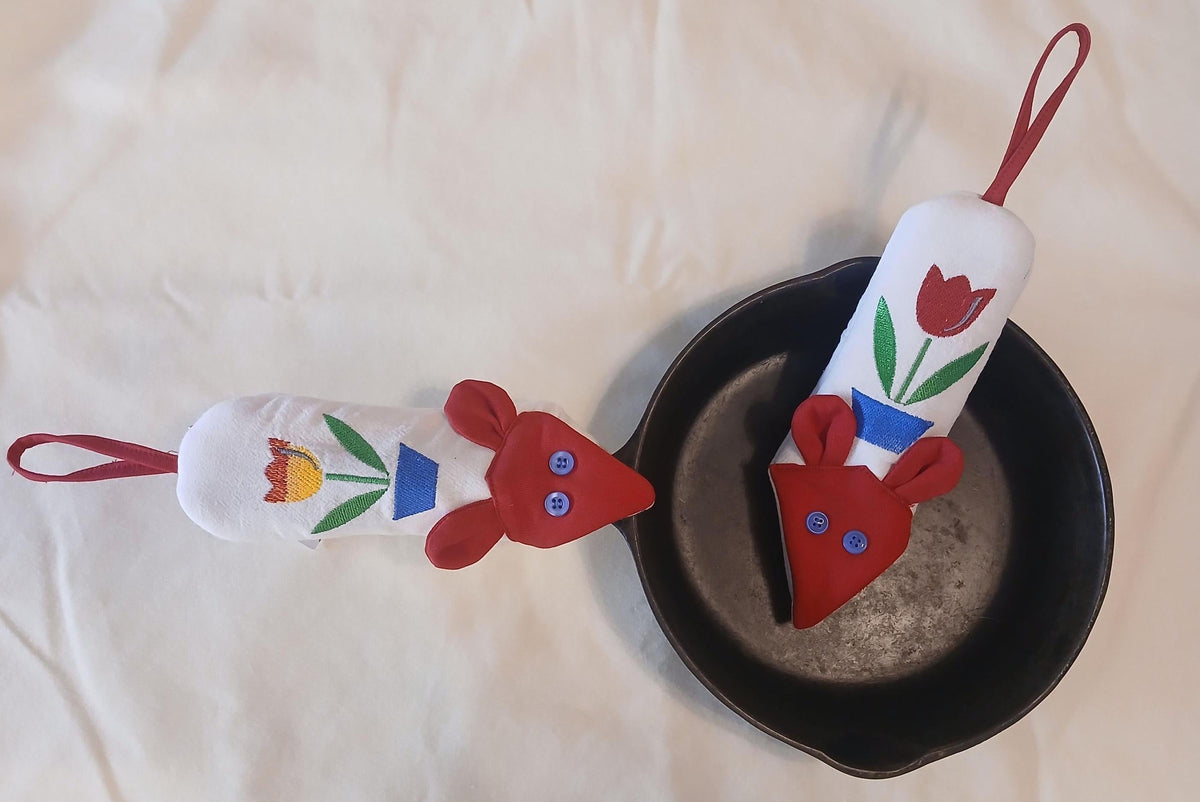 Two white fabric skillet handle potholders, with red faces and colorful floral designs, one resting on a black cast iron skillet and the other on a white fabric background.