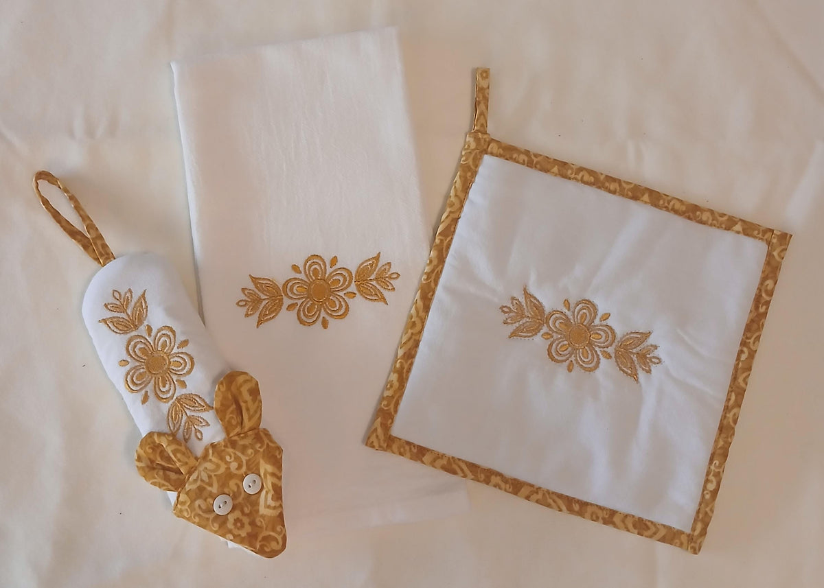 A three-piece kitchen textile set on a cream fabric background, including a white kitchen towel with gold floral embroidery, a potholder with a gold patterned border and matching floral motif, and a handle potholder with a gold patterned face and embroidered design.
