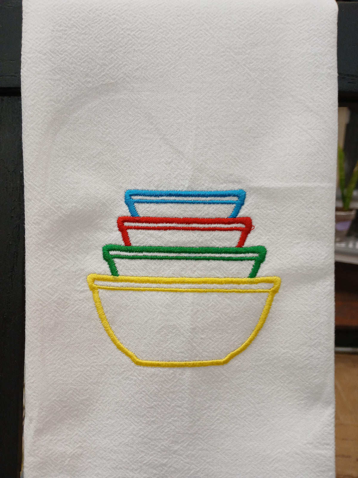 A white kitchen towel featuring an embroidered design of four nested bowls in yellow, green, red, and blue.