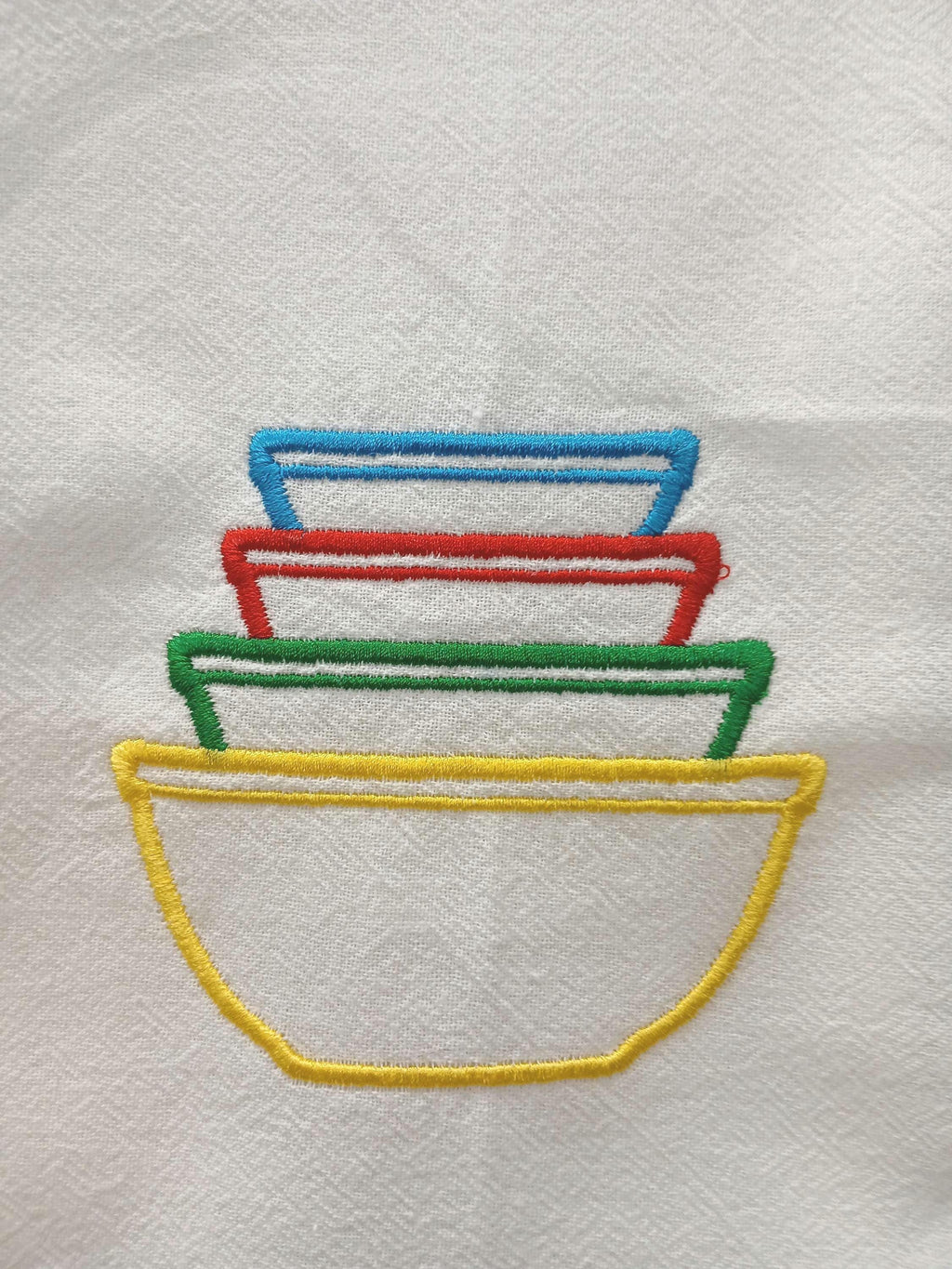 A close-up of embroidered stacked bowls in yellow, green, red, and blue on white fabric.