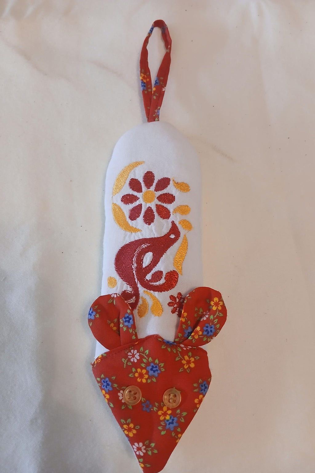 A handmade embroidered fabric handle potholder shaped like a stylized mouse, featuring a white body with red and yellow embroidery, a red floral hanging loop, and a red floral  face with button eyes and fabric ears.