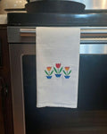 A white kitchen towel hanging from the handle of a stainless steel oven, featuring a small embroidered design of three potted tulips in red, orange, yellow, and blue.
