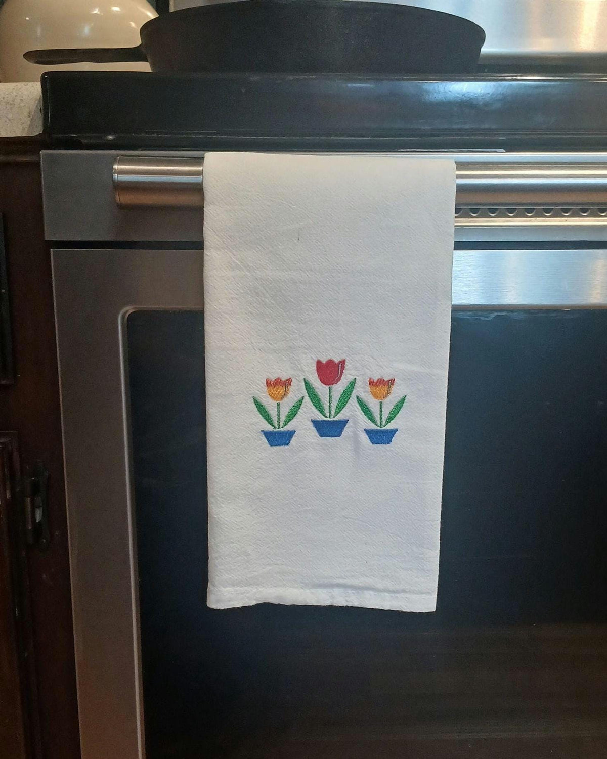 A white kitchen towel hanging from the handle of a stainless steel oven, featuring a small embroidered design of three potted tulips in red, orange, yellow, and blue.