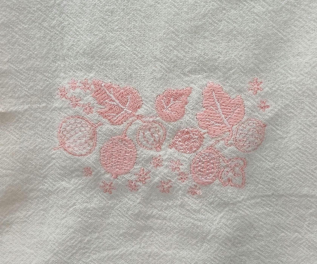 A close-up of a white flour sack towel, featuring a light pink embroidered pattern of leaves, berries, and small floral motifs.