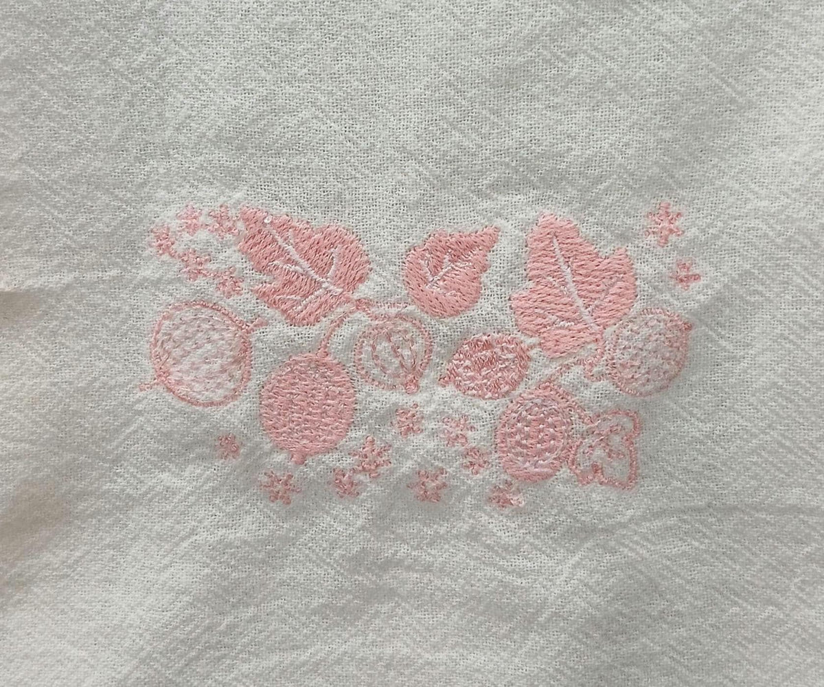 A close-up of a white flour sack towel, featuring a light pink embroidered pattern of leaves, berries, and small floral motifs.