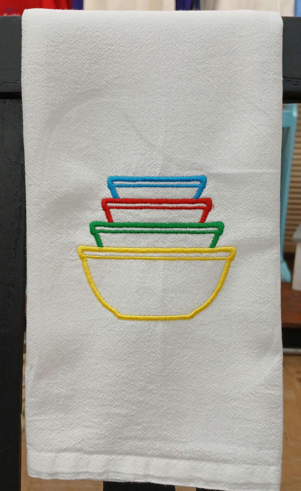 A white kitchen towel featuring an embroidered design of four nested bowls in yellow, green, red, and blue, hanging on a dark rack.