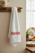 A white kitchen towel with a horizontal embroidered pattern of red birds, orange flowers, and yellow leaves, hanging against a dark background.