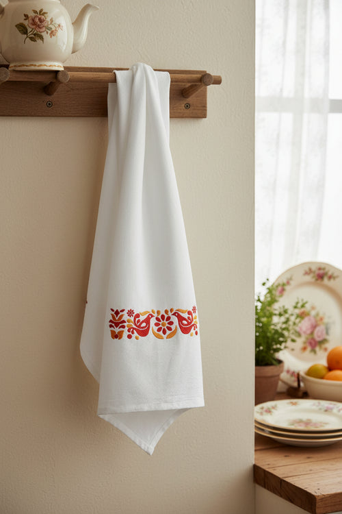 A white kitchen towel with a horizontal embroidered pattern of red birds, orange flowers, and yellow leaves, hanging against a dark background.