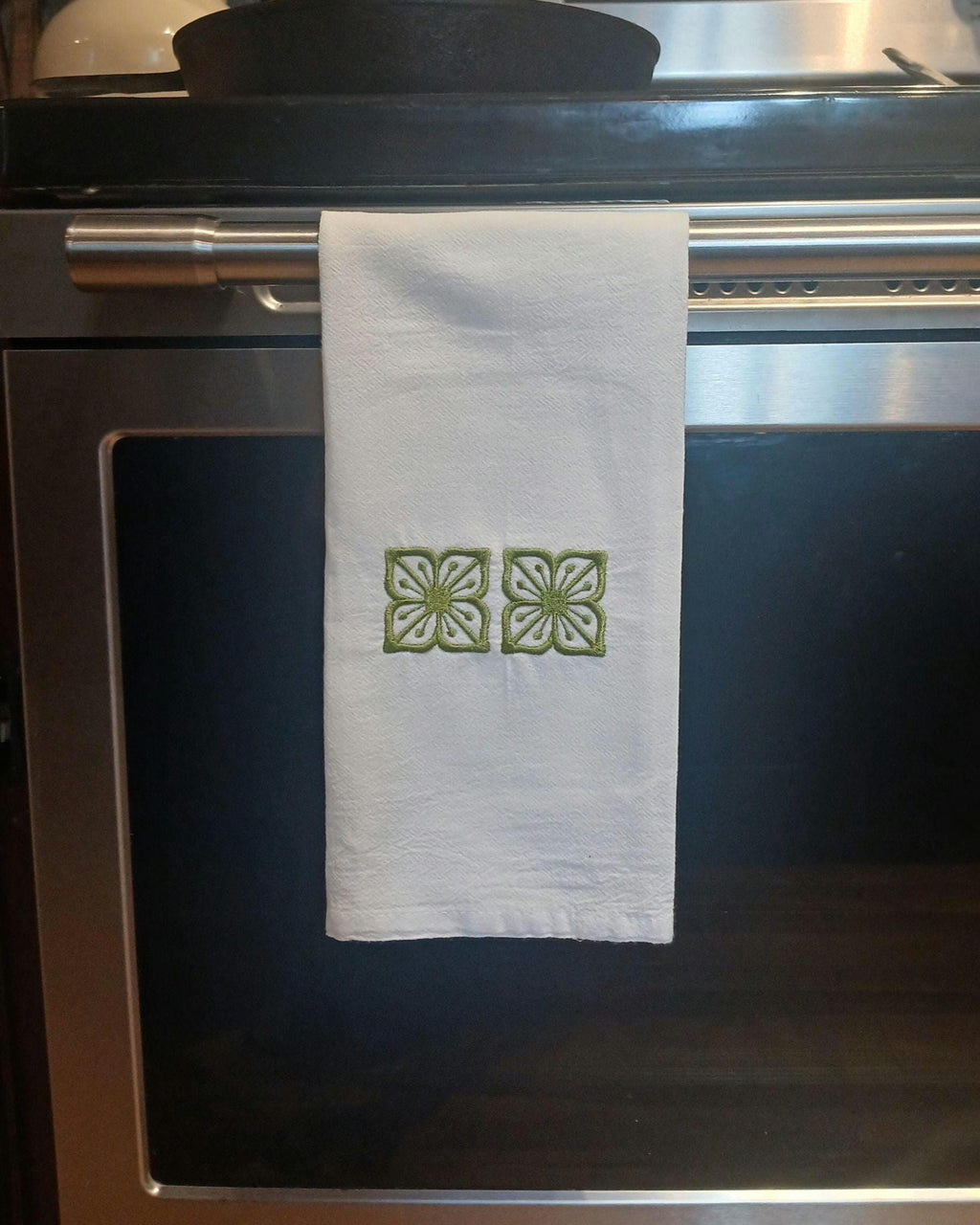 A white kitchen towel with two green embroidered floral motifs hangs from a stainless steel oven handle in a kitchen setting.