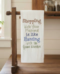 A white fabric kitchen towel hanging from a dark wooden rack, featuring embroidered text that reads 'Shopping with Your Husband is like Hunting with the Game Warden' in brown, green, and blue lettering.