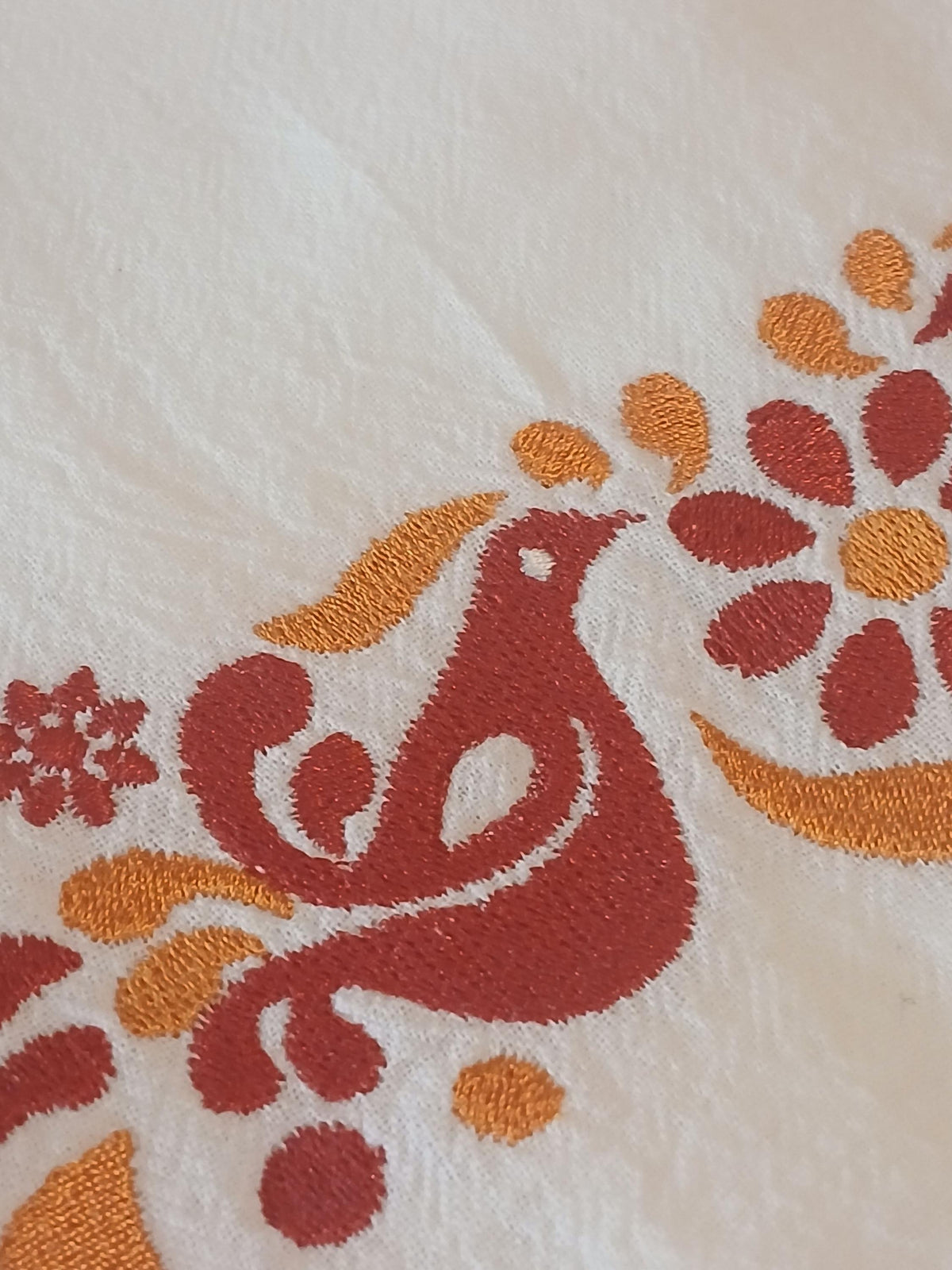 A close-up of a white embroidered textile featuring a stylized bird and floral motifs in rust red and orange.
