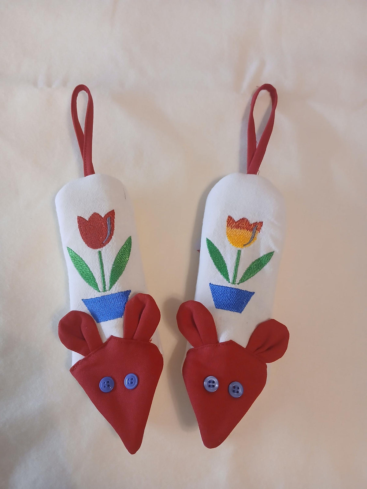 A pair of handmade mouse-shaped handle potholders with red hanging loops, decorated with embroidered tulips, blue button eyes and red ears at the bottom, displayed on a light beige fabric background.