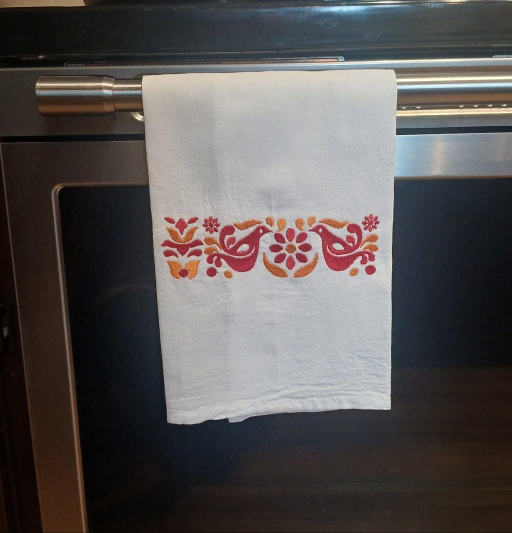 A white kitchen towel with a colorful embroidered floral and bird pattern hangs from the handle of a stainless steel oven in a kitchen setting.