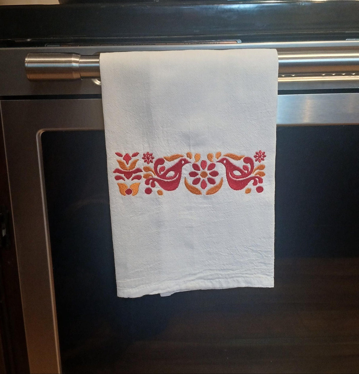 A white kitchen towel with a colorful embroidered floral and bird pattern hangs from the handle of a stainless steel oven in a kitchen setting.