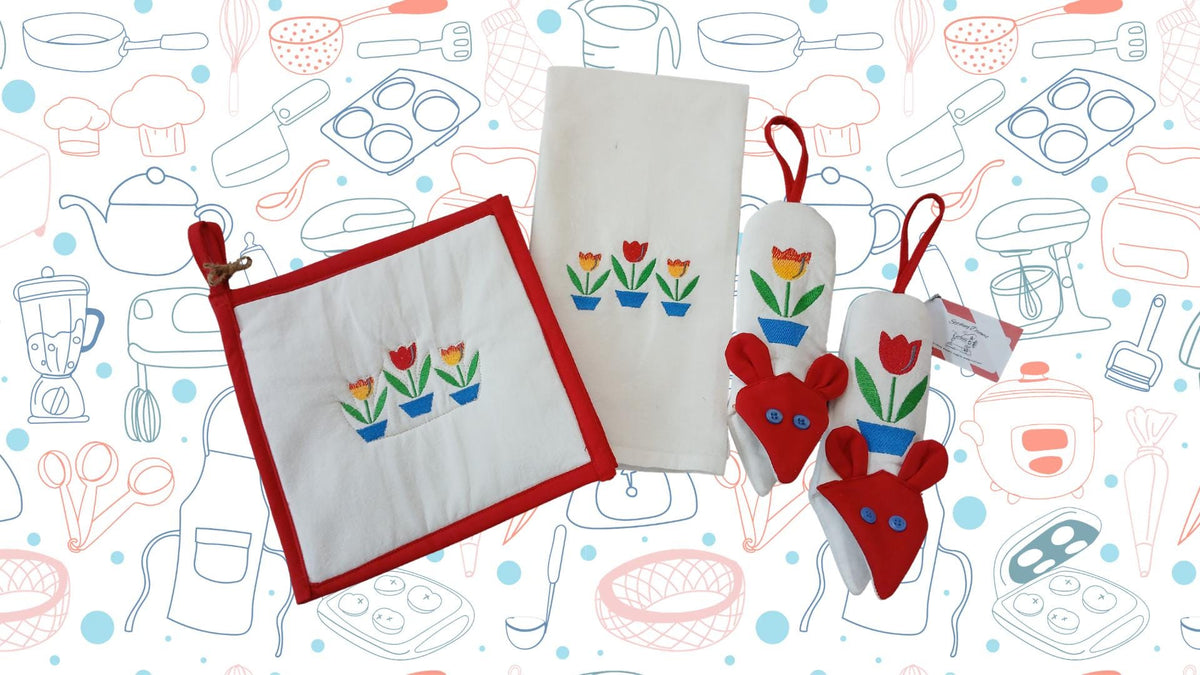 A kitchen accessory set featuring a white potholder with a red border and embroidered tulips, a matching white kitchen towel with the same tulip motif, and a pair of red handle potholders with matching tulip designs, arranged on a patterned background.