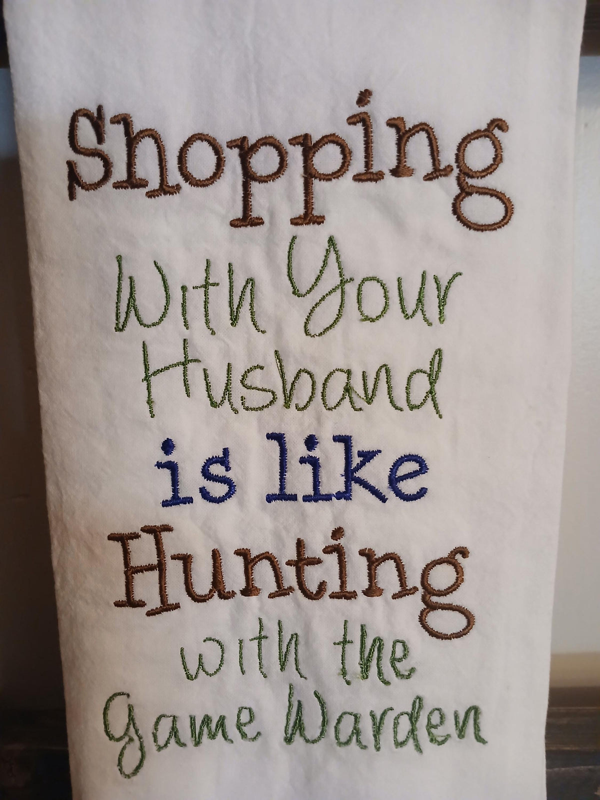 A close-up of a white embroidered towel featuring the humorous phrase 'Shopping with Your Husband is like Hunting with the Game Warden' in brown, green, and blue stitched lettering.