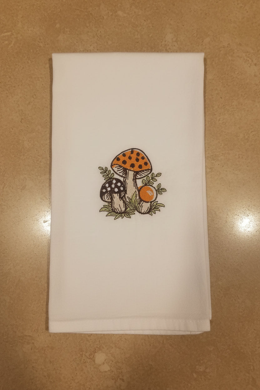A white fabric kitchen towel featuring an embroidered design of three mushrooms (one orange-capped, one black-and-white spotted, and one small orange berry) on a tan background.