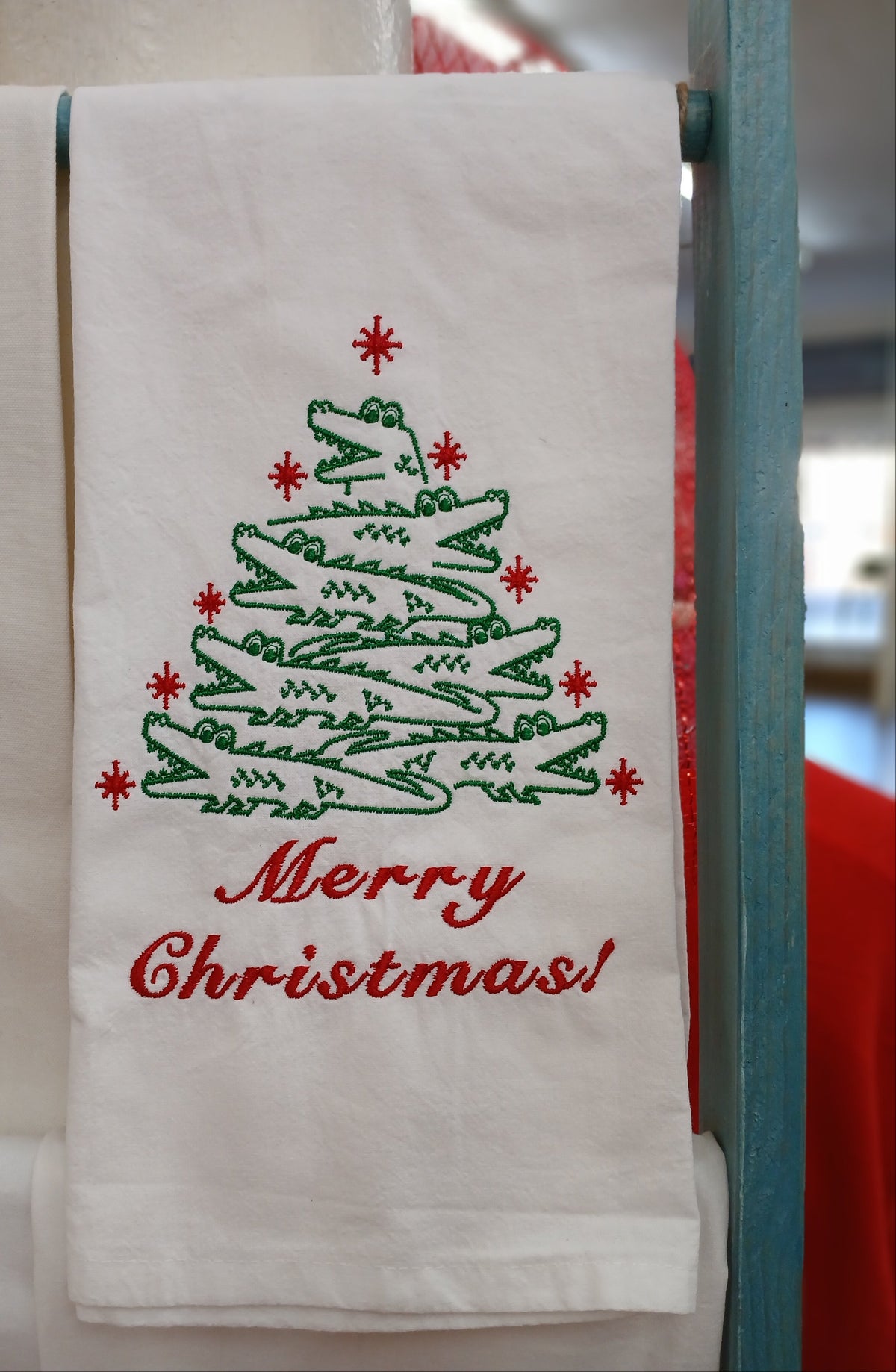 A white kitchen towel with a green alligator tree design and red 'Merry Christmas!' text, hanging on a display rack.

