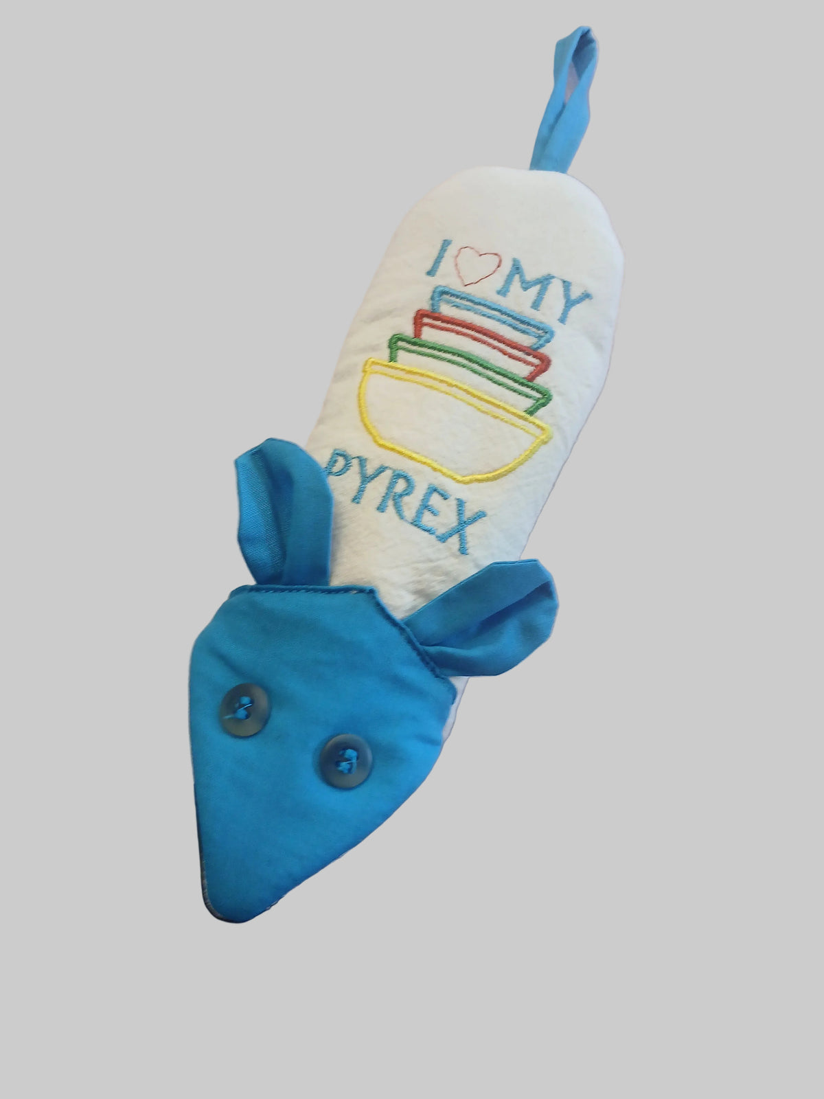 A handmade fabric handle potholder designed to look like a mouse, featuring a blue fabric head with two buttons for eyes and ears, and a white fabric body with embroidered text reading 'I [heart] MY PYREX' and colorful stacked bowls. It is resting on a light beige fabric surface.