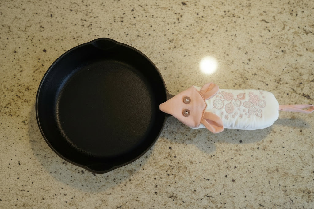 A black cast iron skillet with a pink and white mouse-shaped handle potholder, laid flat on a tan granite countertop.