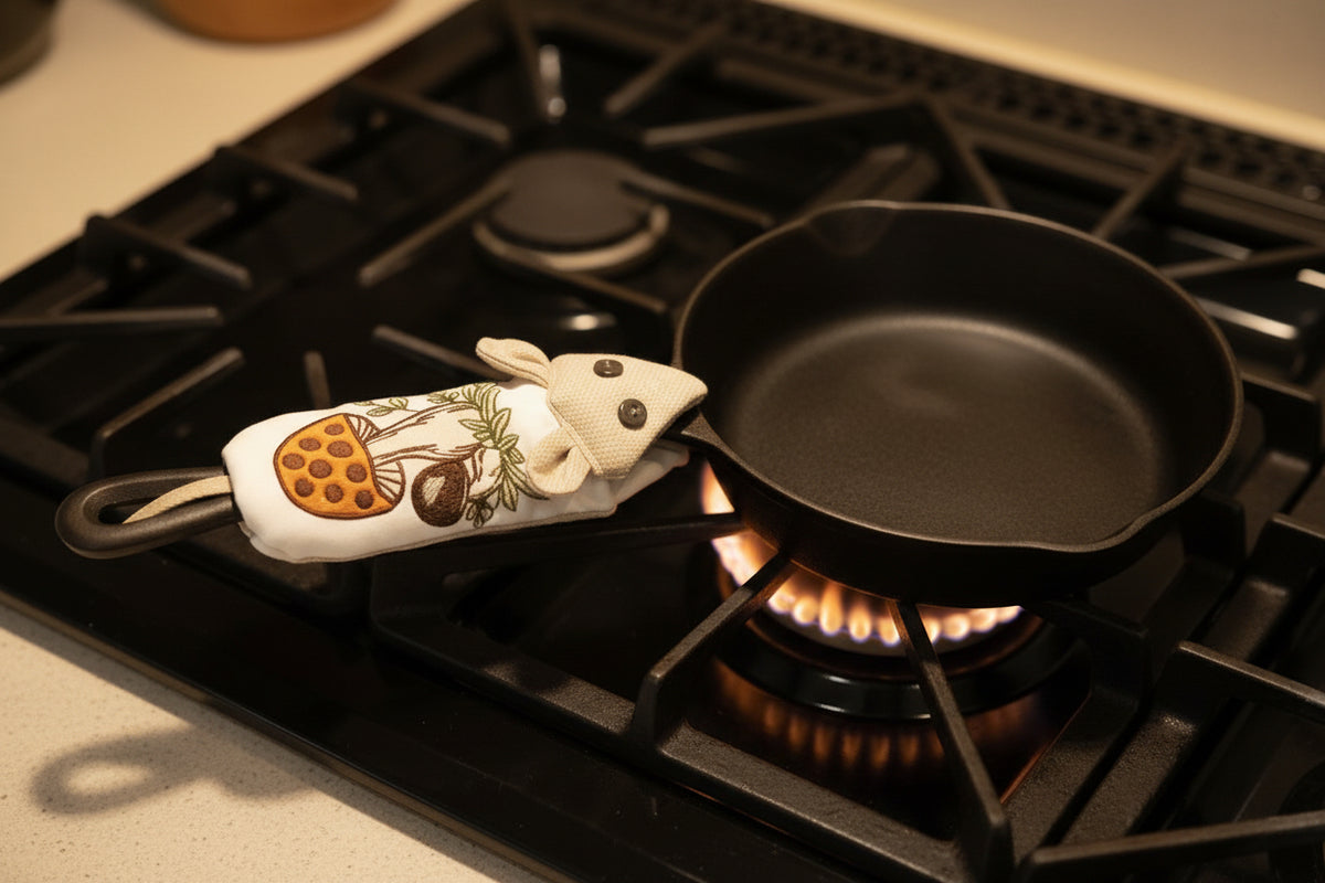 A small black cast-iron skillet with a handle potholder, featuring an embroidered mushroom design, placed on a stove top.