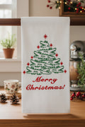 A white kitchen towel with a green alligator tree and red 'Merry Christmas!' text, hanging in a kitchen setting.