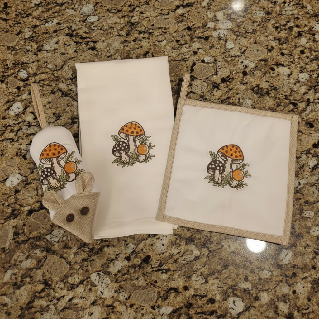 Three cream-colored fabric kitchen accessories with an embroidered mushroom design, laid out on a tan countertop. The set includes a small mouse-shaped handle potholder, rectangular potholder, and a kitchen towel.