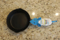 A black cast iron skillet with a blue and white handle potholder featuring the text 'I <3 MY PYREX', displayed on a tan surface.