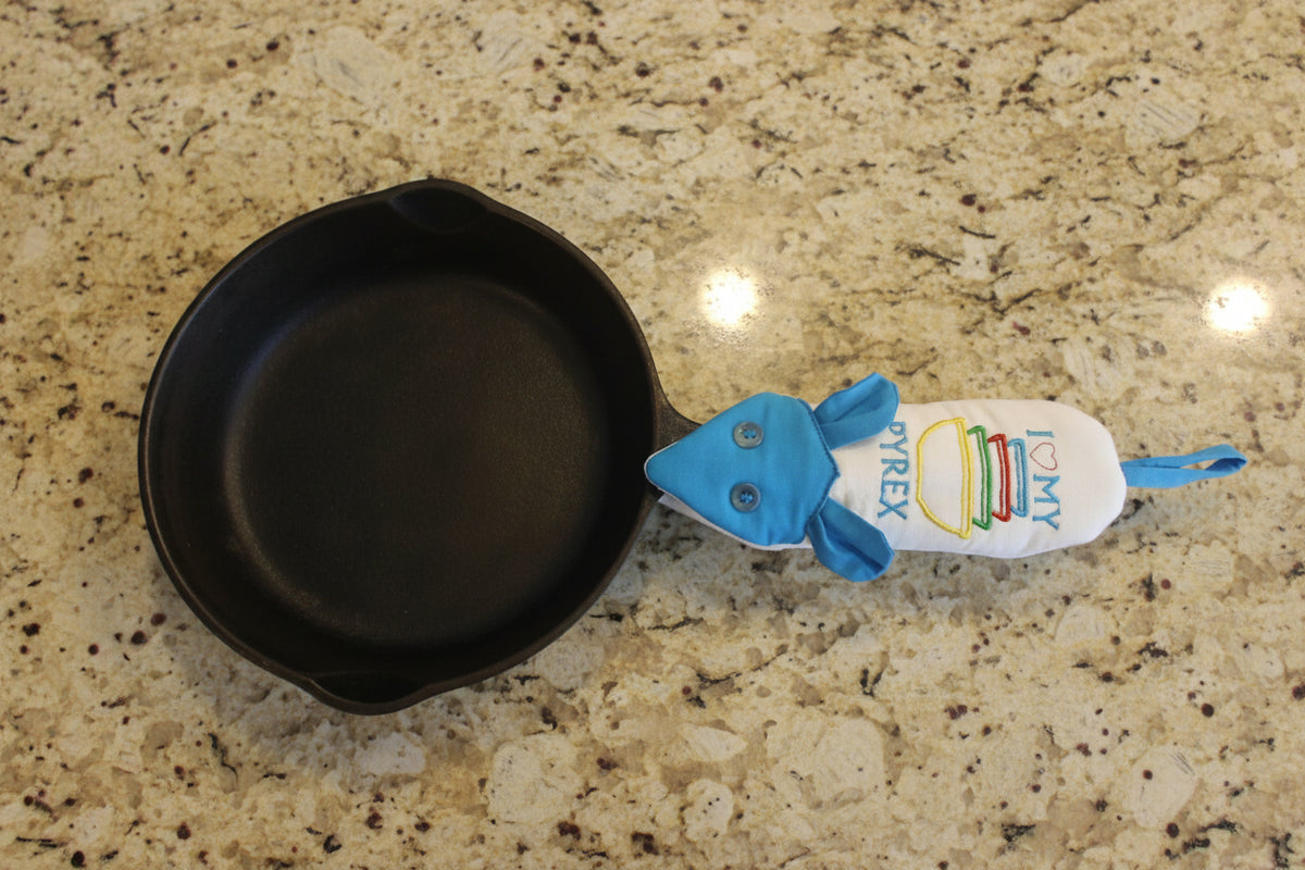 A black cast iron skillet with a blue and white handle potholder featuring the text 'I <3 MY PYREX', displayed on a tan surface.
