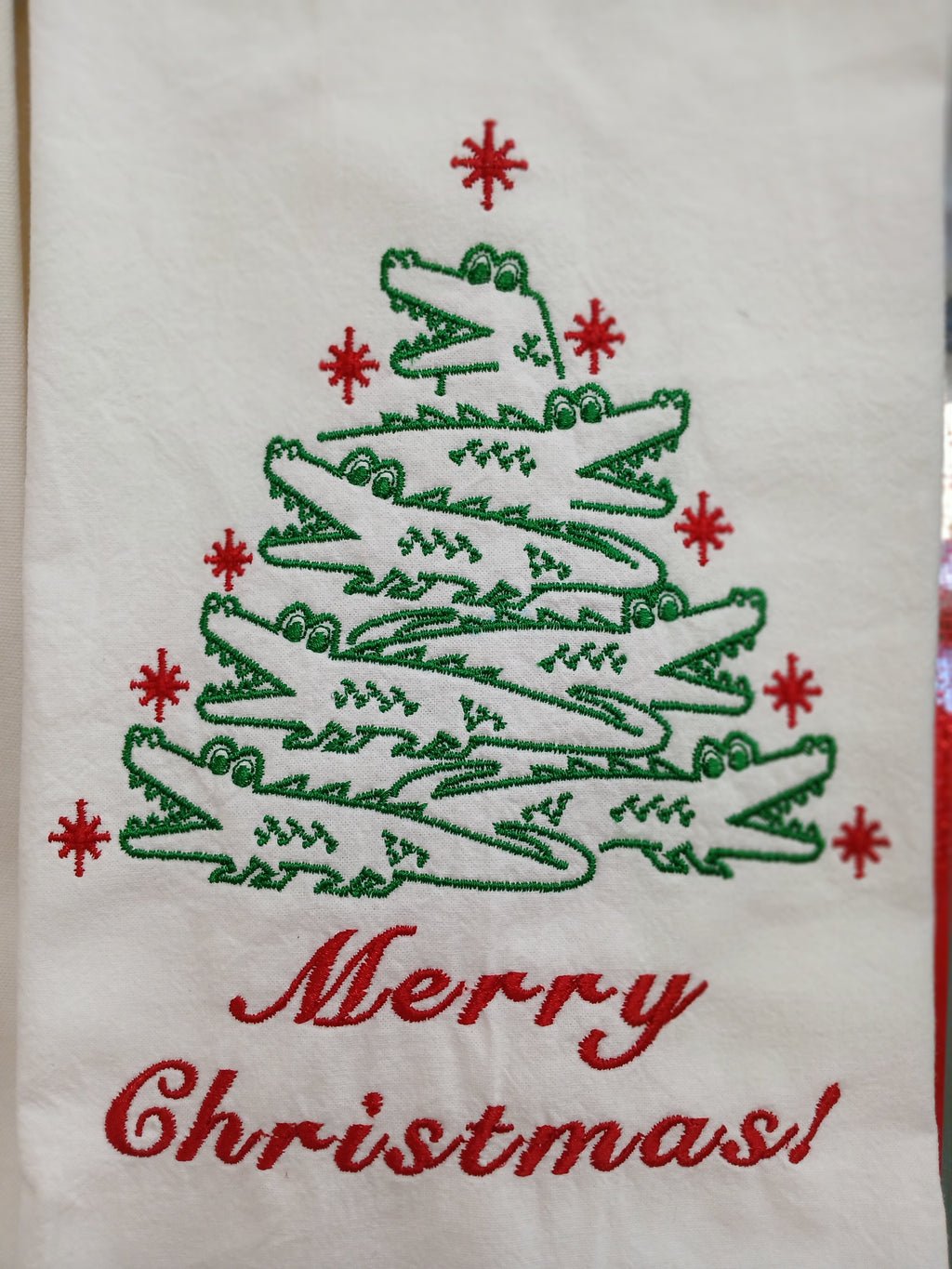 An embroidered Christmas towel featuring a tree made of green alligators, red snowflake accents, and the words 'Merry Christmas!' in red script on a white fabric background.